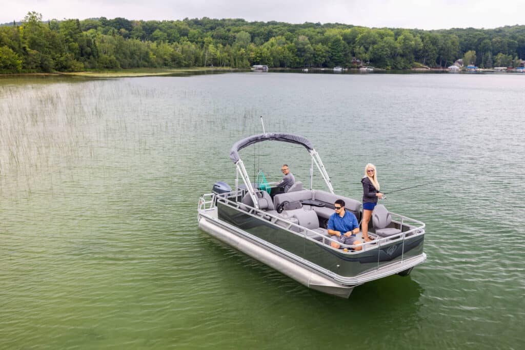 Three people are on a pontoon boat on a lake. One person is seated at the controls, another person is sitting at the front with a fishing rod, and a third person is standing while fishing. The lake is surrounded by dense green trees.