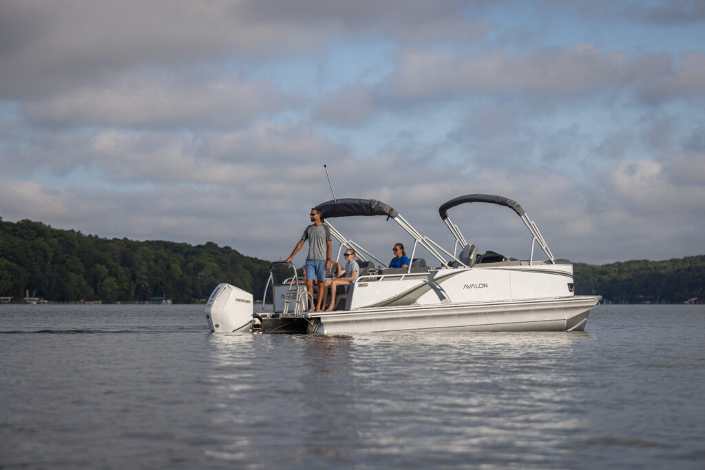 A silver pontoon boat with three people onboard is cruising on a calm lake. The sky is partly cloudy, and dense green trees line the distant shoreline. The group appears relaxed, enjoying the serene water setting.