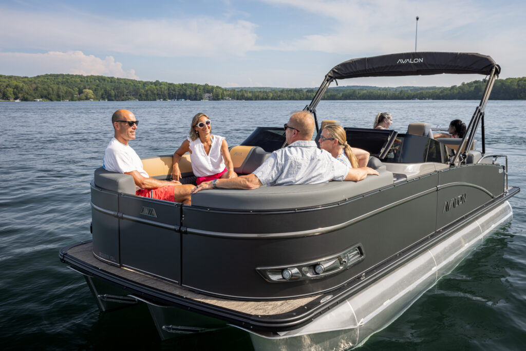 Four people are sitting and chatting on a Catalina Quad Lounger Shift Windshield pontoon boat in calm lake waters. The boat's canopy is partially extended, and green forested hills line the distant shore under a clear blue sky.
